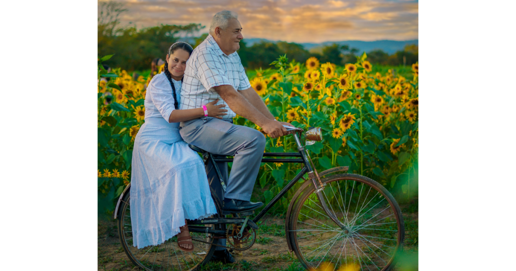 An elderly couple on a bicycle, with the husband carrying his wife, passing in front of a sunflower field with a beautiful sunset.